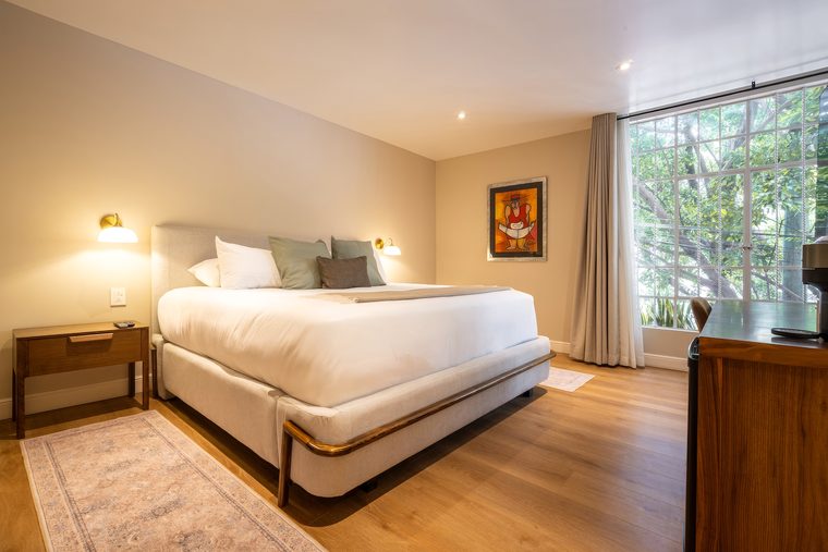 Cama king vestida en blanco, con ventanal a la calle arbolada y un cuadro pequeño sobre la cabecera
