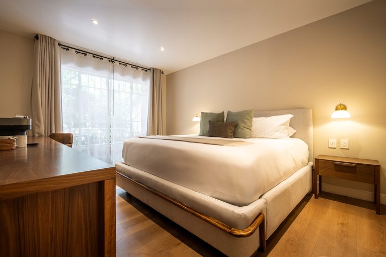 Cama king con cojines verdes junto a la ventana, con una lámpara de pared encendida