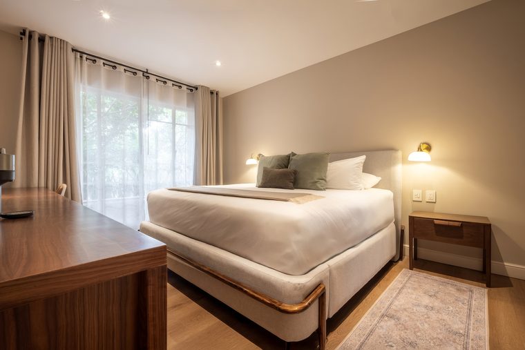 Cama king junto a la ventana, con lámpara de pared y buró de madera